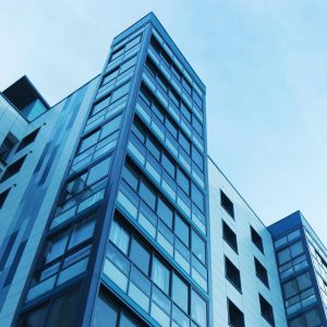 Upward view of a blue-tinted modern glass office building with many windows and sharp angles against a clear sky.