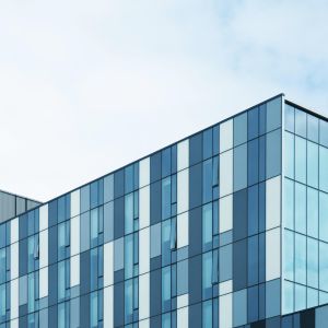 Modern glass office building with blue-tinted panels along a diagonal corner against a pale sky.