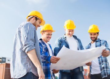 Four construction workers in yellow hard hats study a large blueprint together outdoors at a construction site.