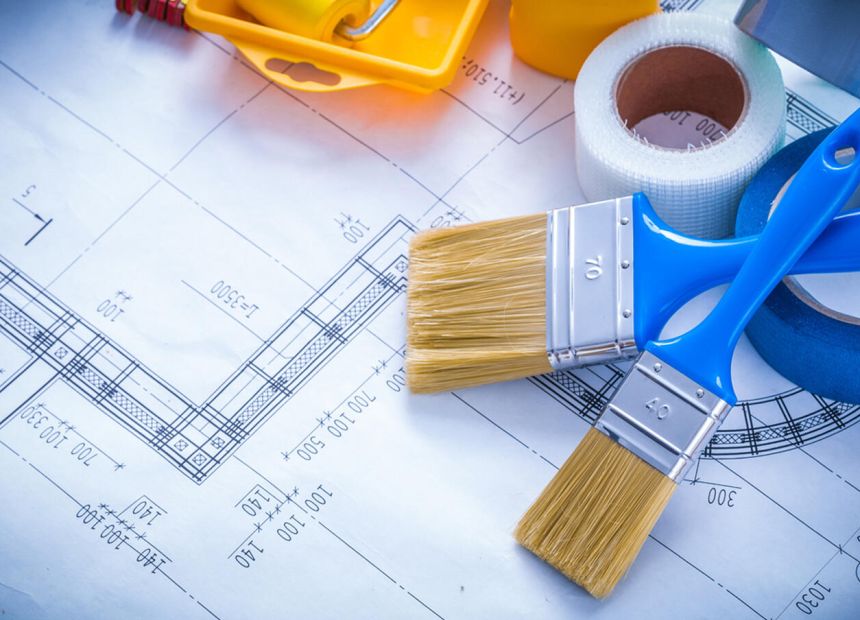 Architectural blueprints with painting supplies (brushes, masking tape, and a tape measure) laid out on the plans to suggest renovation work.